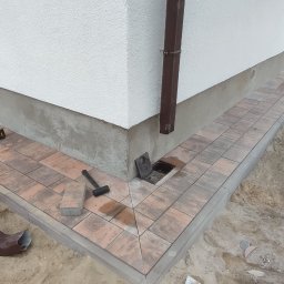 BRUK-TOK MATEUSZ TOKARSKI - Układanie kostki brukowej wokół domu, widoczny fragment nowej ścieżki z kostki w odcieniach brązu, narzędzia pracy: młotek, wkrętarka, oraz karton i rura spustowa w tle, teren wokół w trakcie prac...