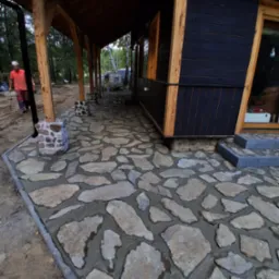 Kamienny chodnik z nieregularnych płyt prowadzący wzdłuż drewnianego domu z czarną elewacją. Widoczny fragment ogrodu i pracownik w oddali.