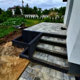 Schody i taras wyłożone szarą kostką brukową, z elementami czarnego obramowania. Widok na ogród i dom w tle. Ukończona realizacja z dbałością o detale.