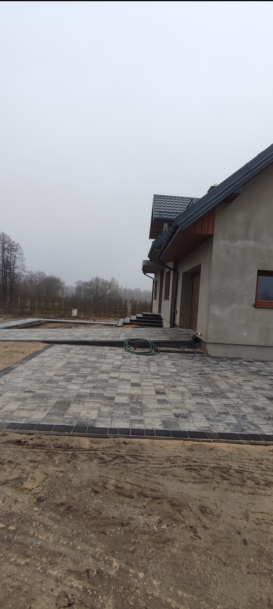 Ułożona kostka brukowa przed domem jednorodzinnym w pochmurny dzień, widoczny zwinięty wąż ogrodowy na kostce.