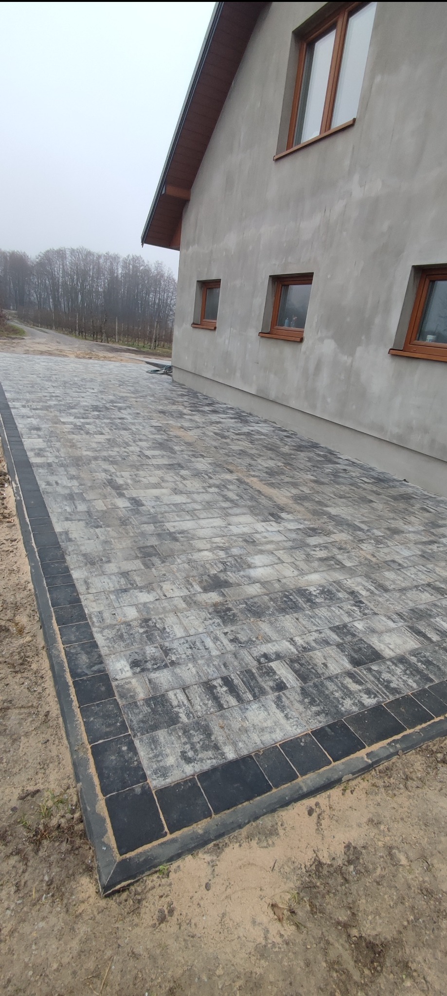 Ułożona kostka brukowa w odcieniach szarości z czarną obwódką przy ścianie budynku z trzema oknami, widoczny fragment drogi i drzewa w tle.