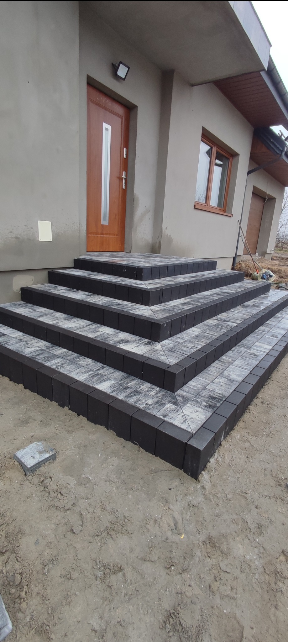 Schody wejściowe do domu wyłożone kostką brukową w odcieniach szarości i czerni, widoczne niedociągnięcia w postaci pojedynczej kostki leżącej obok oraz brudne podłoże.
