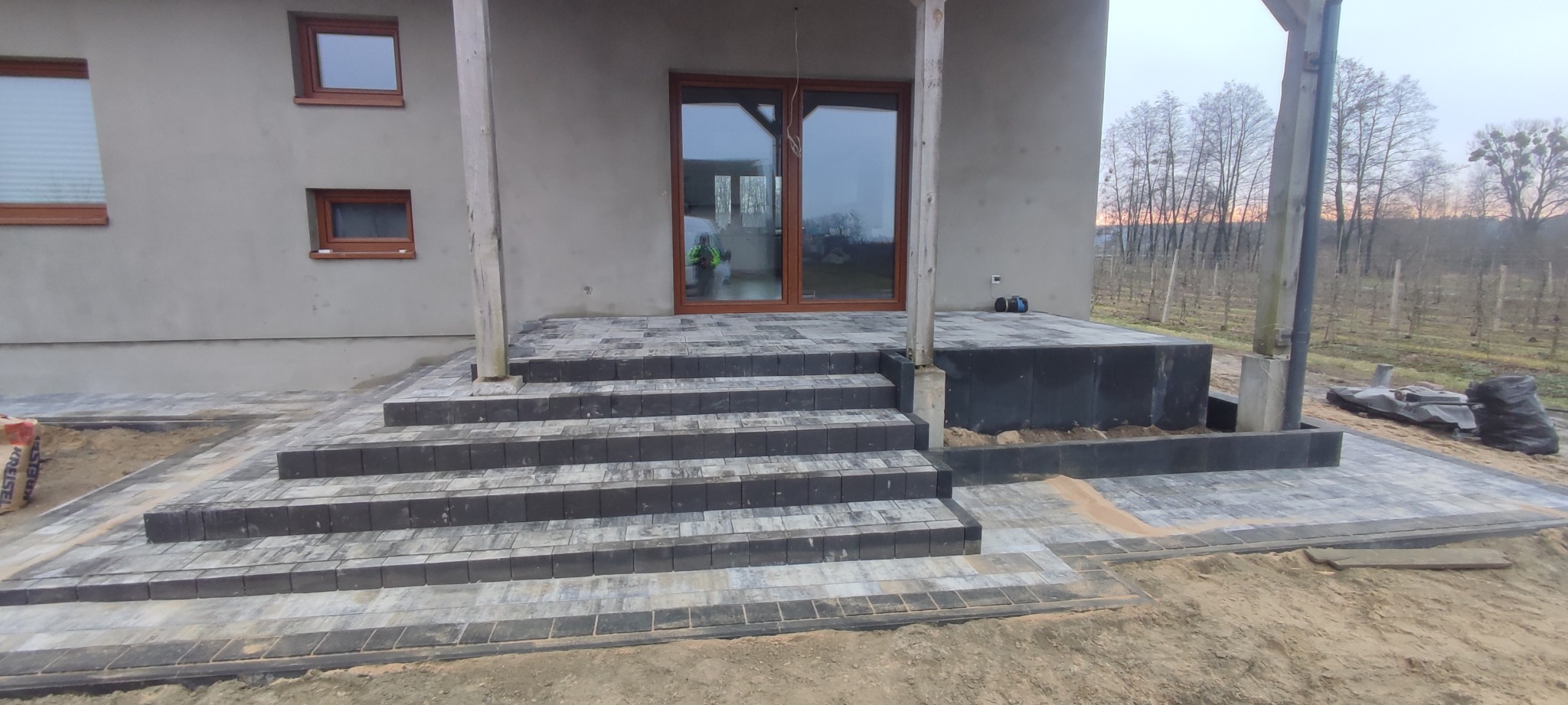 Montaż schodów z kostki brukowej prowadzących do domu z drewnianą stolarką okienną, widoczne worki z zaprawą i niezakończone prace ziemne.