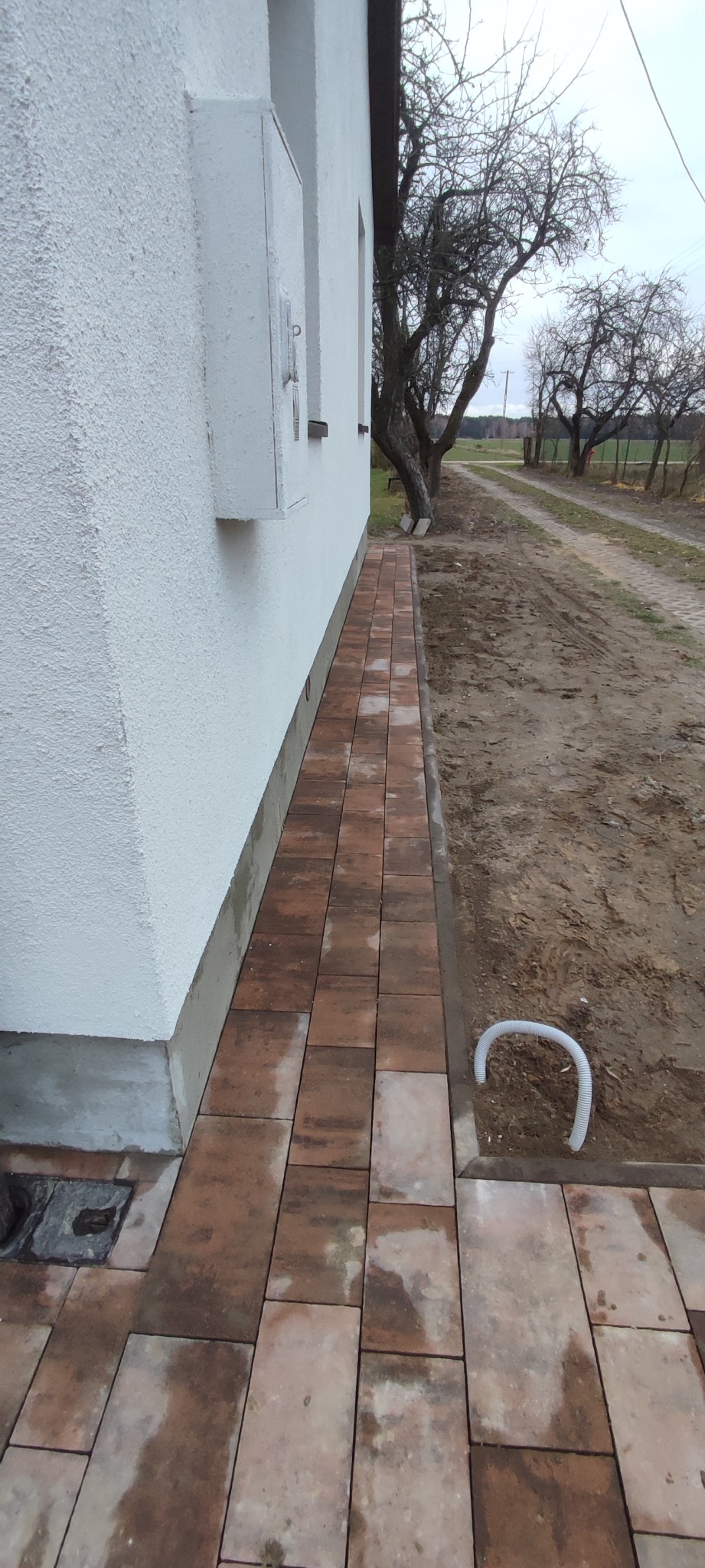 Ułożona ścieżka z kostki w odcieniach czerwieni i brązu, przylegająca do białej ściany budynku, z widoczną rurą w ziemi i drzewami w tle.