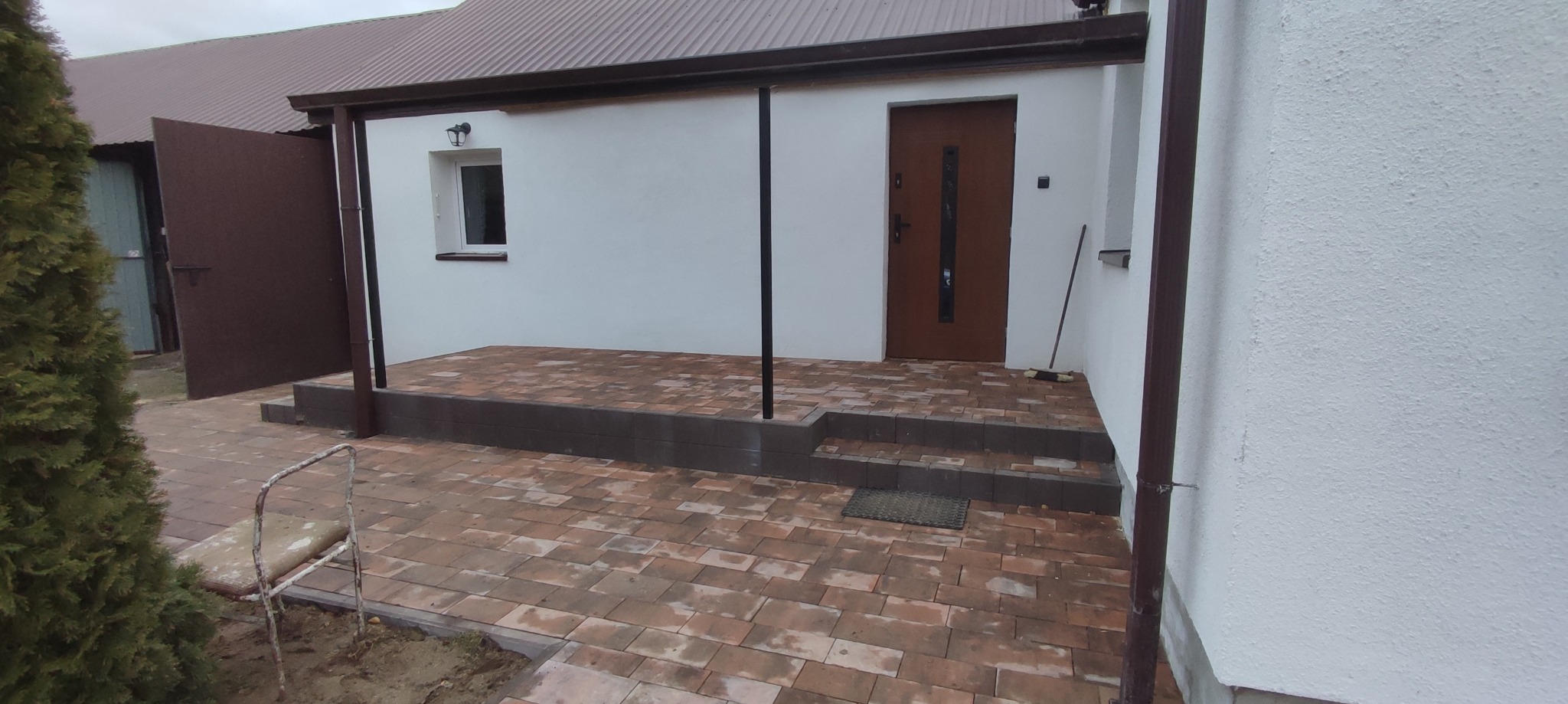 Wykończony taras z kostki brukowej w odcieniach brązu i czerwieni, prowadzący do wejścia do budynku z brązowymi drzwiami; widoczny fragment ogrodu z tujami po lewej stronie.
