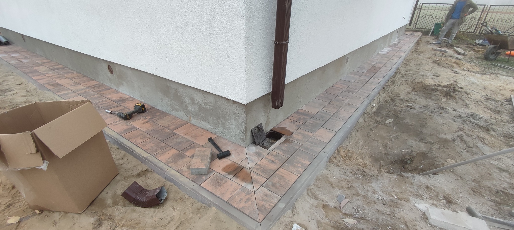 Układanie kostki brukowej wokół domu, widoczny fragment nowej ścieżki z kostki w odcieniach brązu, narzędzia pracy: młotek, wkrętarka, oraz karton i rura spustowa w tle, teren wokół w trakcie prac...