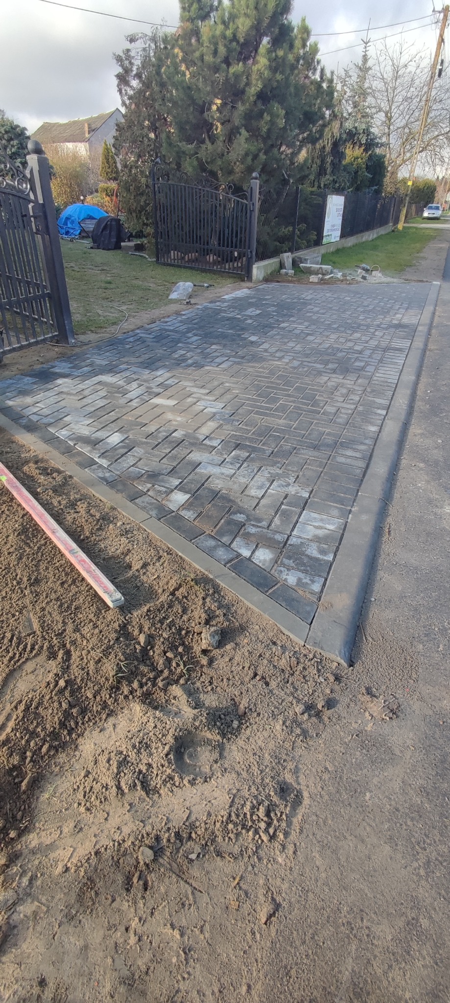 Ułożona nawierzchnia z kostki brukowej w szaro-grafitowej tonacji, obramowana ciemniejszymi obrzeżami, widoczny poziomica i przygotowane podłoże z piasku, brama w tle.