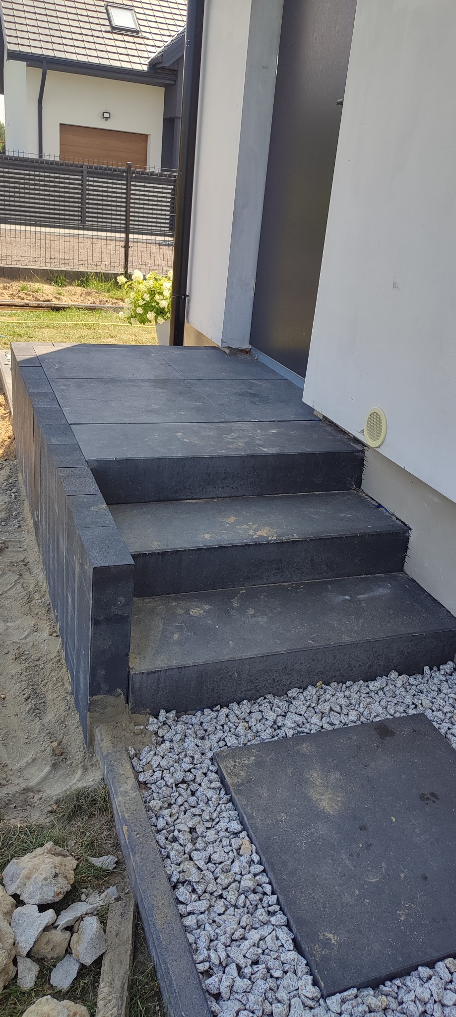 Schody wejściowe do domu wyłożone ciemną kostką brukową, obrzeża z kostki, wysypane ozdobnym grysem, widok na fragment elewacji budynku i ogrodzenie.