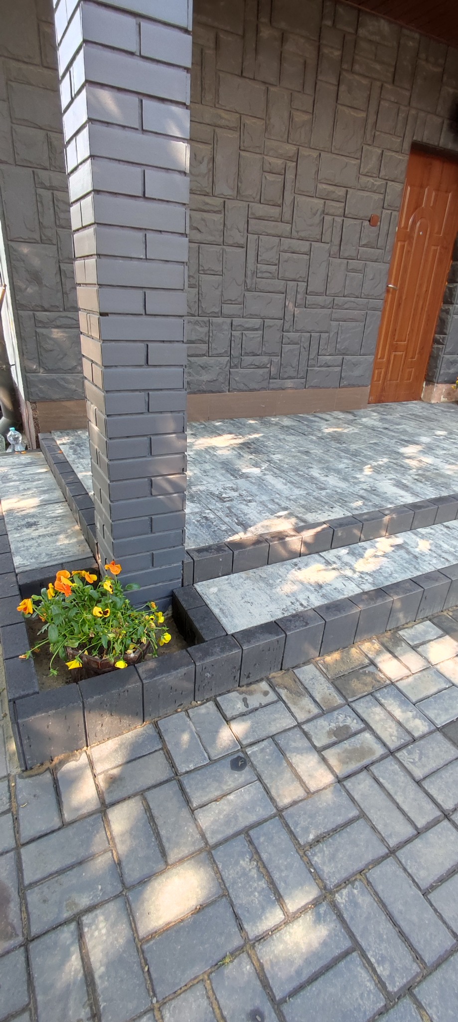 Szara kostka brukowa ułożona w jodełkę, z obrzeżami i kolumną z cegły klinkierowej, donica z kwiatami u podstawy kolumny, elewacja budynku z szarej cegły dekoracyjnej, brązowe drzwi wejściowe.