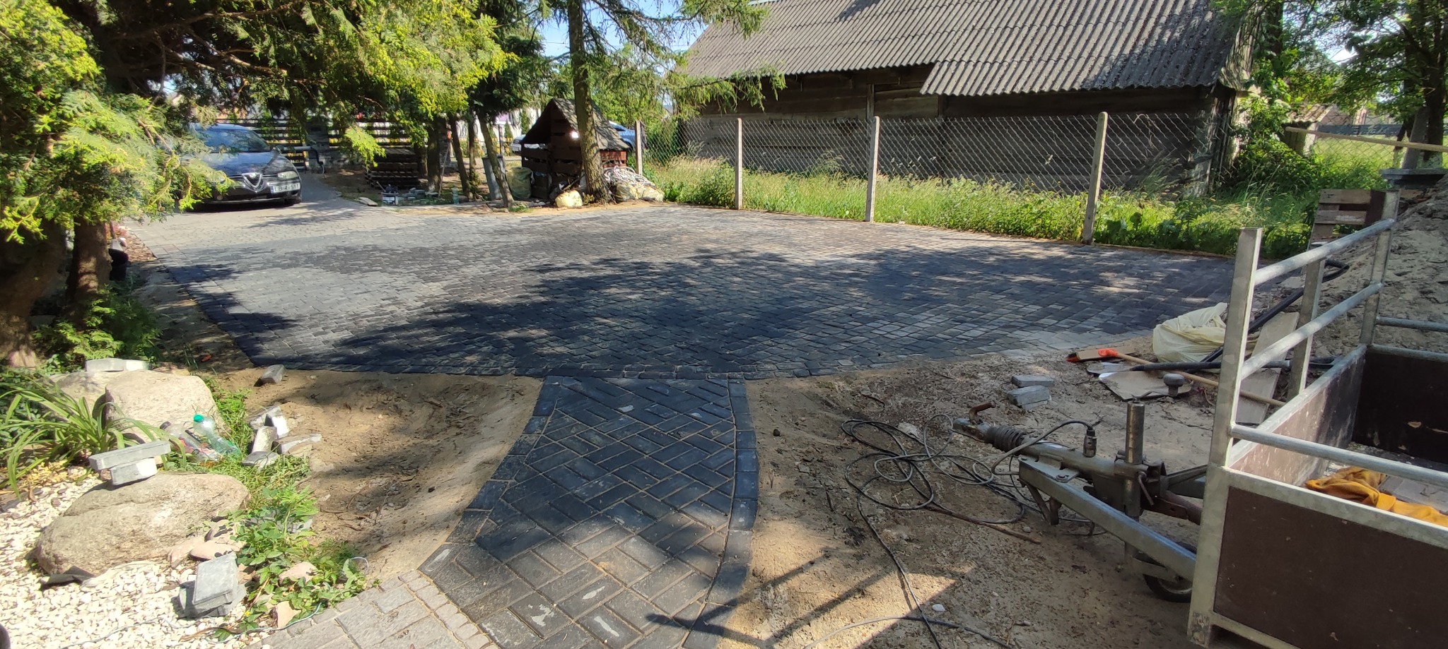 Ułożona nawierzchnia z kostki brukowej w odcieniach szarości, widoczny fragment terenu przygotowanego do dalszych prac, przyczepa i narzędzia budowlane w tle, drzewa i budynki w oddali.