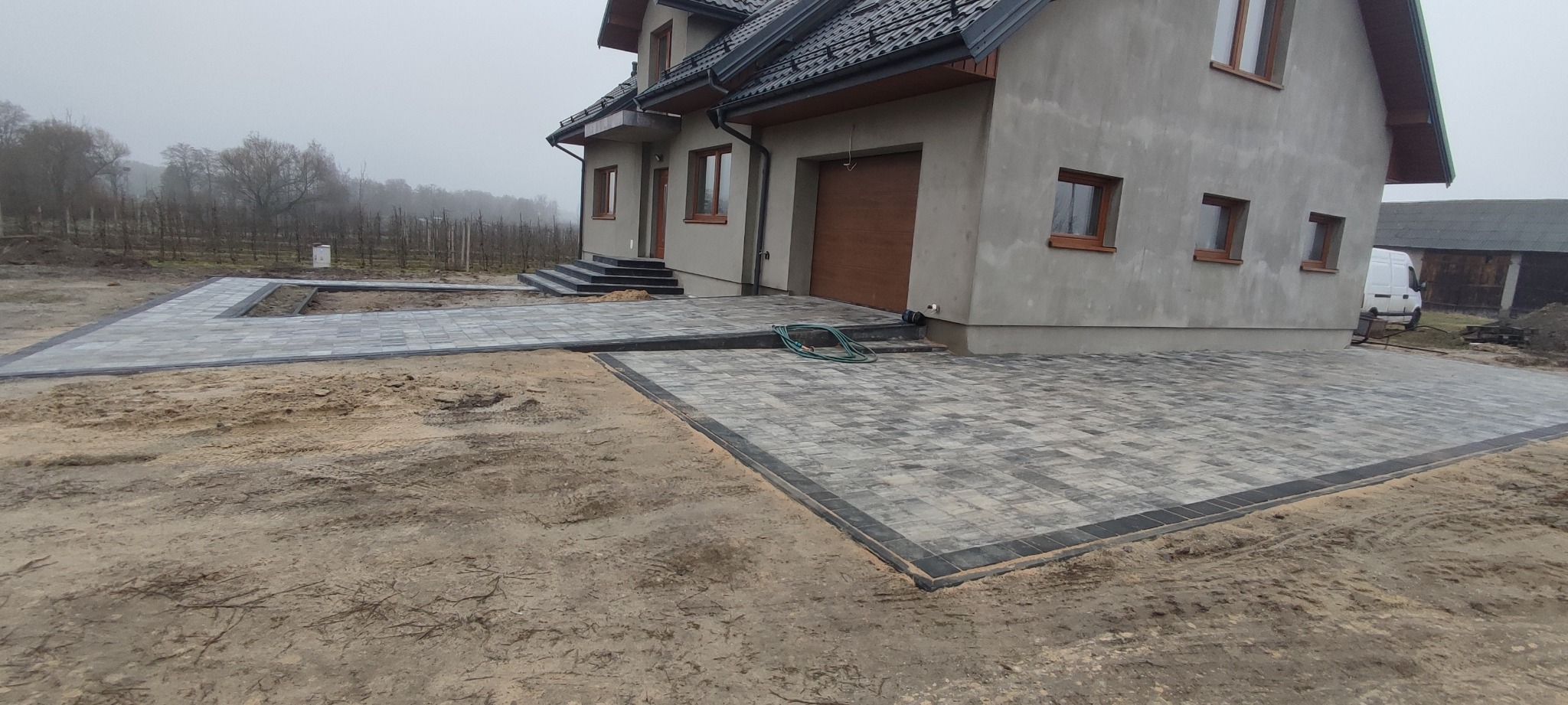 Ułożona szara kostka brukowa z obramowaniem w kolorze antracytu tworząca podjazd i chodnik przed domem z garażem; widoczny zwinięty wąż ogrodowy, trawnik w trakcie zakładania oraz drzewa w tle.