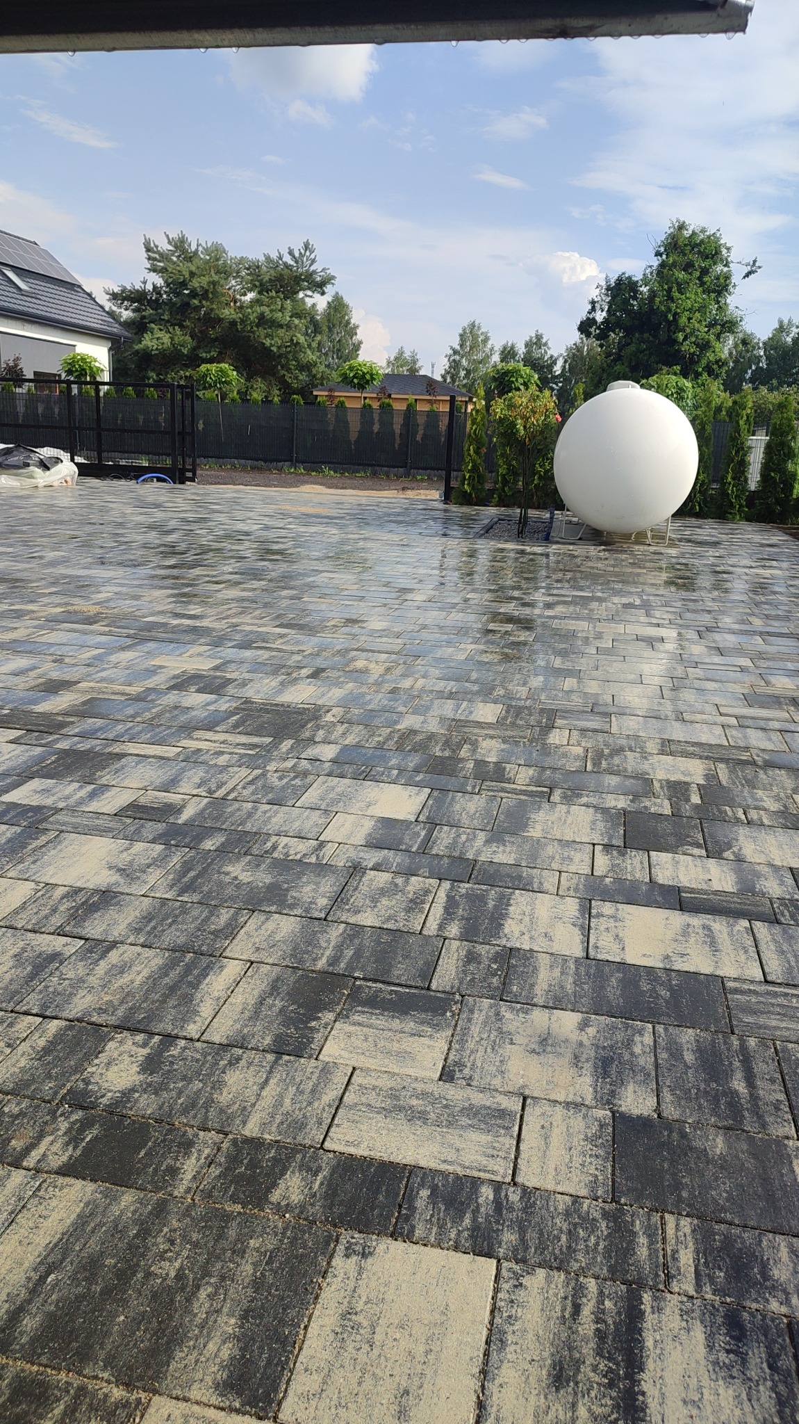 Ułożona, mokra kostka brukowa w odcieniach szarości i beżu, z widocznym zbiornikiem na gaz w tle i fragmentem domu po lewej stronie