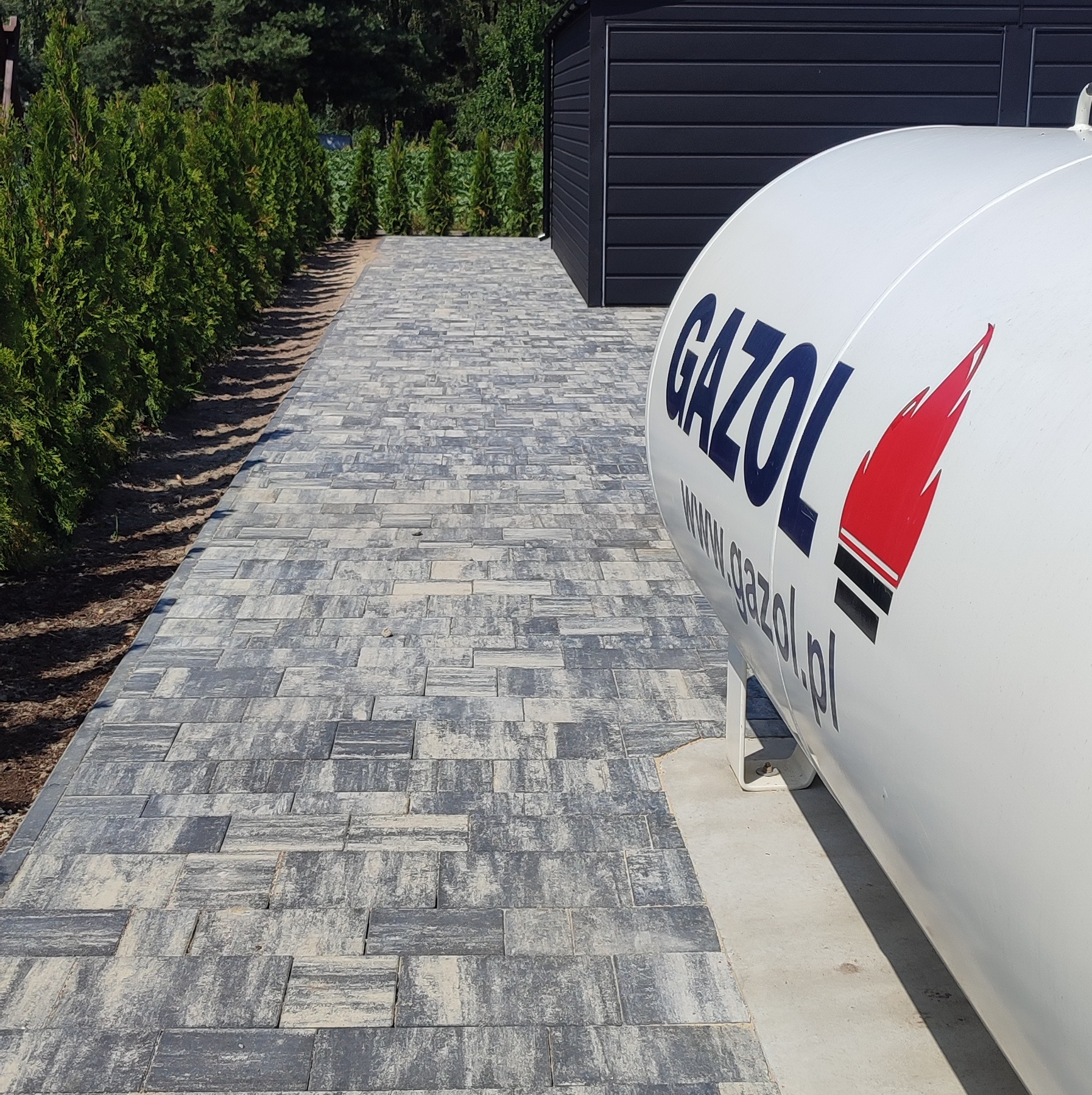 Ułożona kostka brukowa w odcieniach szarości prowadząca do ciemnego domku narzędziowego, z widocznym zbiornikiem gazu GAZOL z boku kadru.