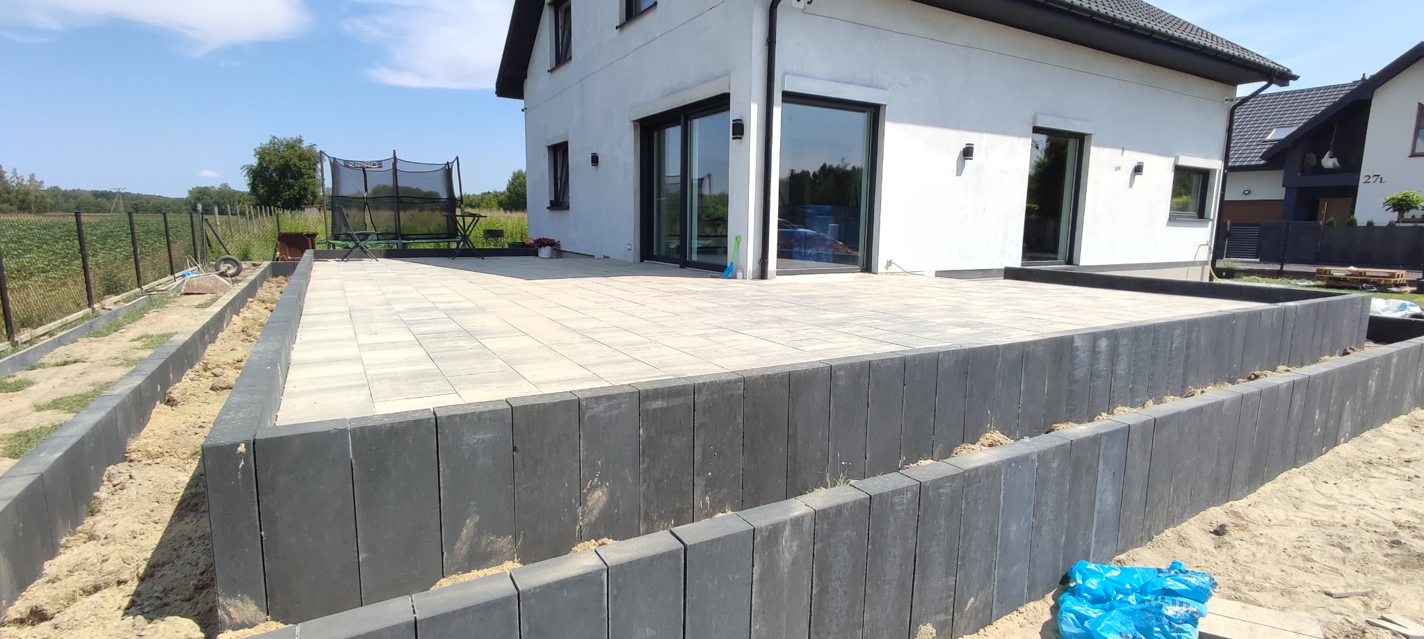 Jasnoszara kostka brukowa ułożona na podwyższonym tarasie przydomowym z ciemnoszarą obudową, widok na ogród i dom z dużymi oknami przesuwnymi.