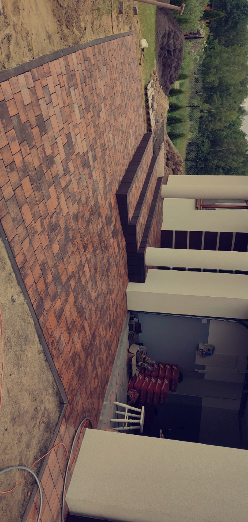 Ułożona kostka brukowa w odcieniach brązu prowadząca do schodów wejściowych budynku, widoczne worki z zaprawą i teren przygotowany pod dalsze prace.