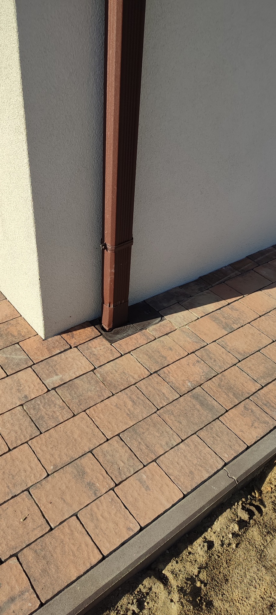 Ułożona kostka brukowa w odcieniach brązu i beżu, z widoczną rynną spustową przy ścianie budynku, obrzeża chodnika z szarego betonu, częściowo obsypane ziemią.