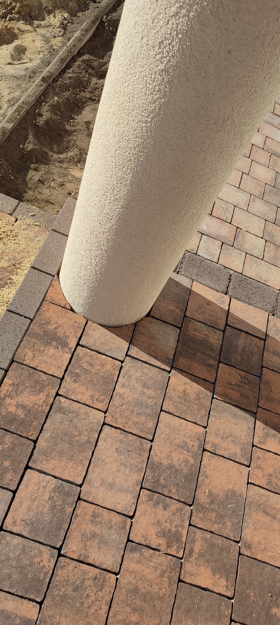 Fragment nowej nawierzchni z brązowej kostki brukowej ułożonej wokół okrągłego, teksturowanego słupa, widoczny wykop pod obrzeże.
