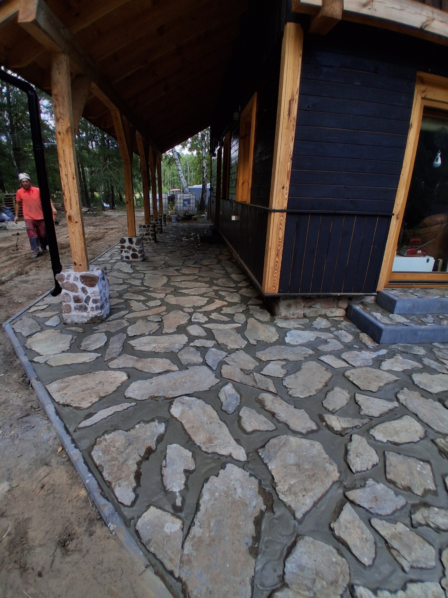 Kamienny chodnik z nieregularnych płyt prowadzący wzdłuż drewnianego domu z czarną elewacją. Widoczny fragment ogrodu i pracownik w oddali.