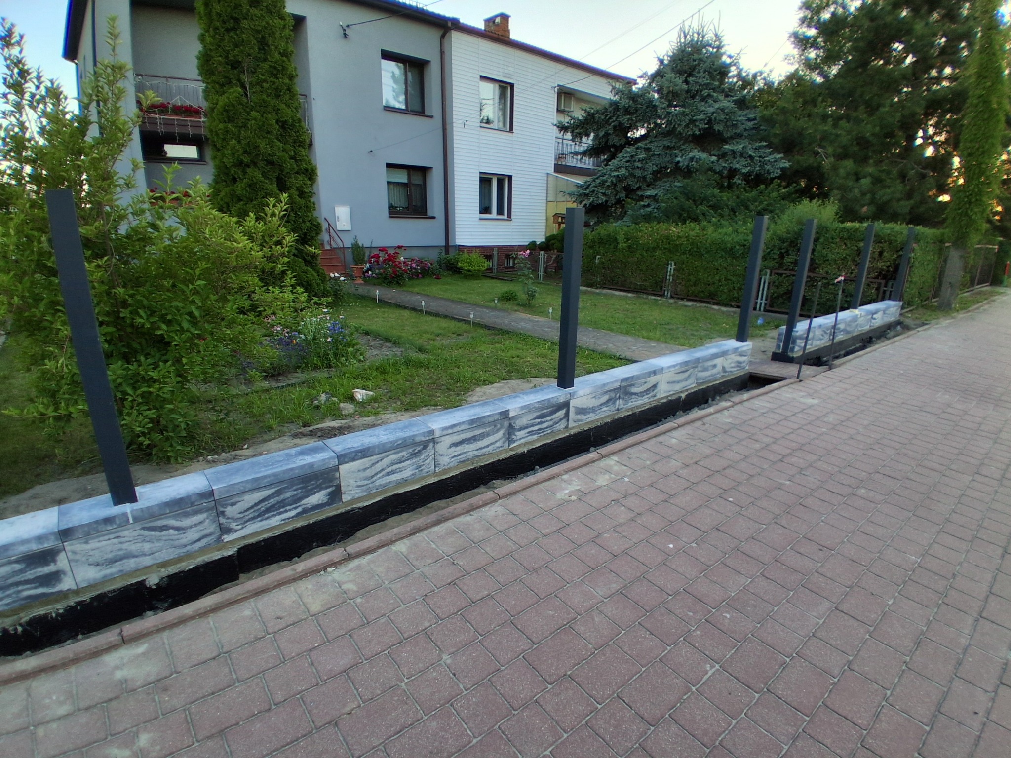 Wyrównane obrzeże z szarej kostki brukowej z czarnymi słupkami ogrodzeniowymi, widok z góry na chodnik z czerwonej kostki, dom i zieleń w tle.