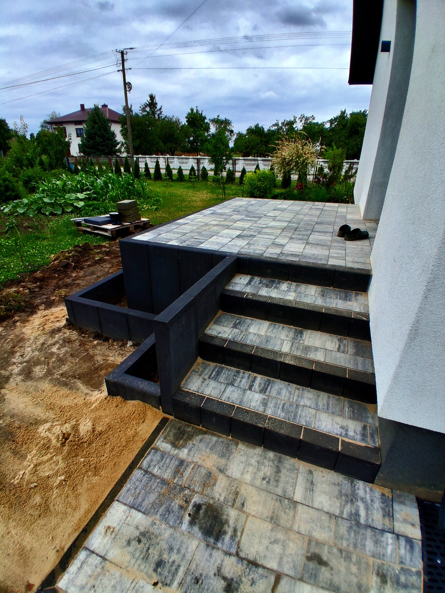 Schody i taras wyłożone szarą kostką brukową, z elementami czarnego obramowania. Widok na ogród i dom w tle. Ukończona realizacja z dbałością o detale.