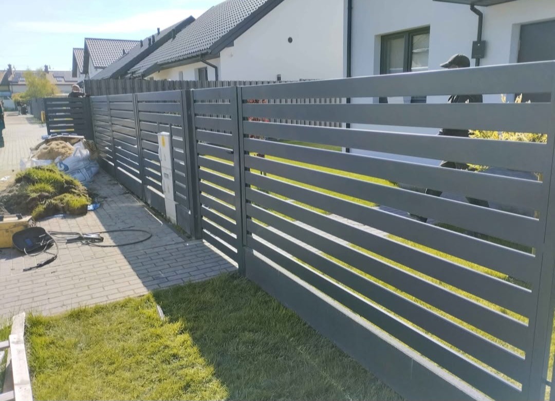Nowoczesne, poziome ogrodzenie panelowe w kolorze grafitowym, oddzielające posesję od chodnika. Widoczne trawnik i fragmenty kostki brukowej. W tle domy jednorodzinne.