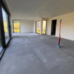 Wnętrze nowo wykończonego domu, widoczne świeżo położone tynki na ścianach i suficie, z dużymi oknami i drzwiami balkonowymi, na podłodze wylewka samopoziomująca.