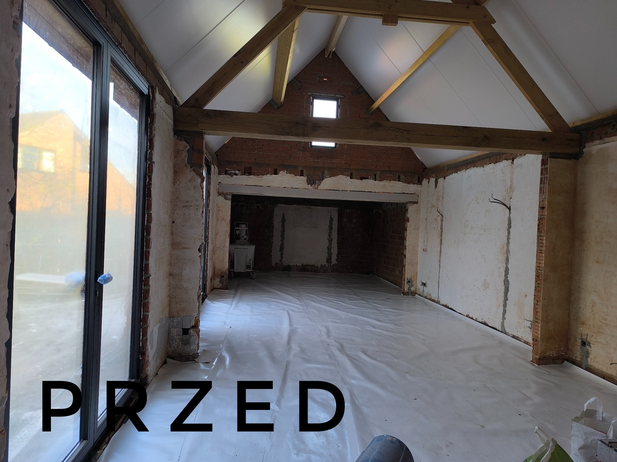 Wnętrze w Świebodzinie przed remontem: surowe ściany z odsłoniętą cegłą, drewniane belki stropowe, podłoga zabezpieczona folią ochronną, widoczne przygotowania do prac wykończeniowych.