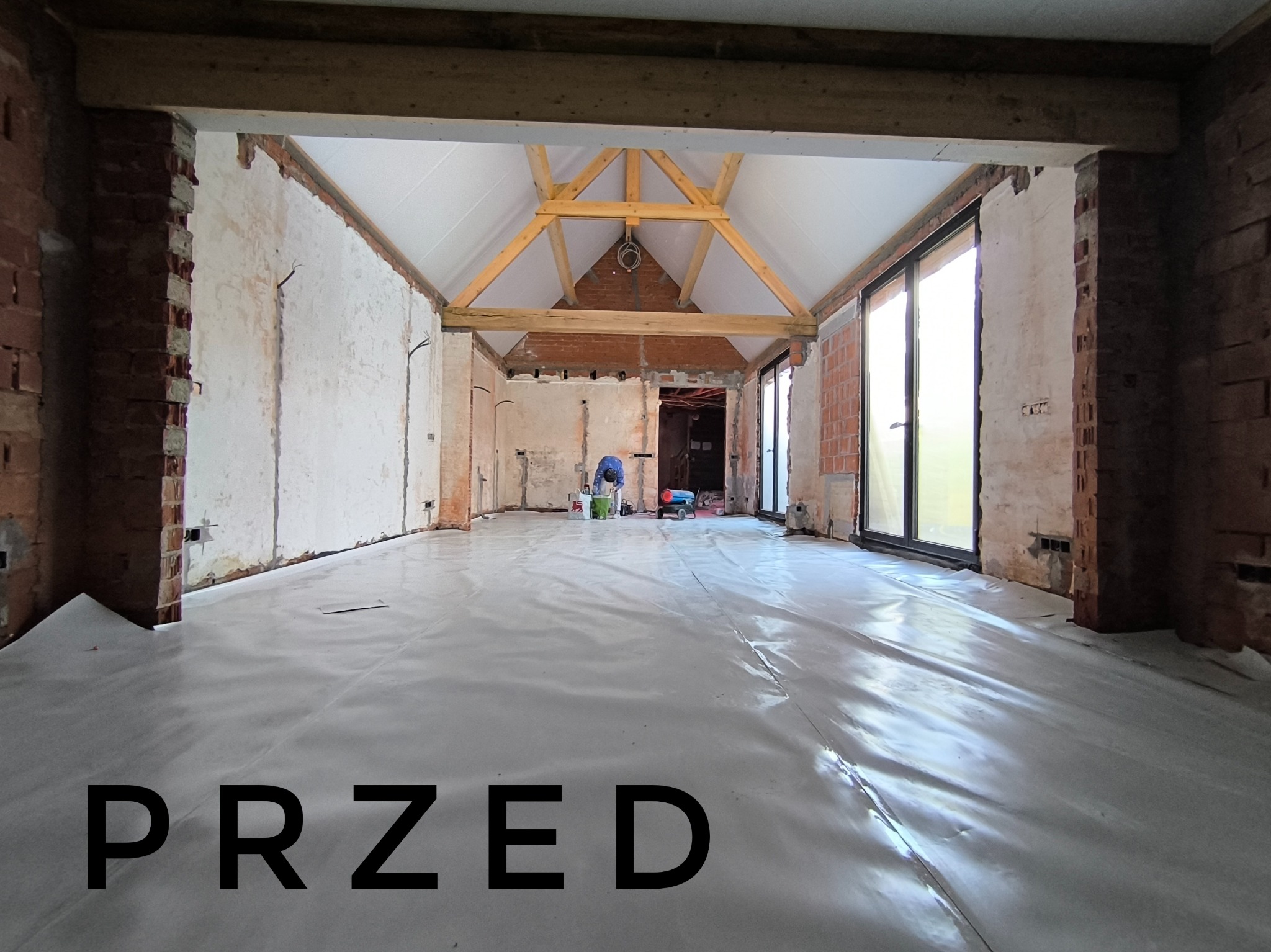 Wnętrze w trakcie remontu: surowe ceglane ściany, drewniana konstrukcja dachu, podłoga zabezpieczona folią, widoczny pracownik przygotowujący się do prac wykończeniowych.