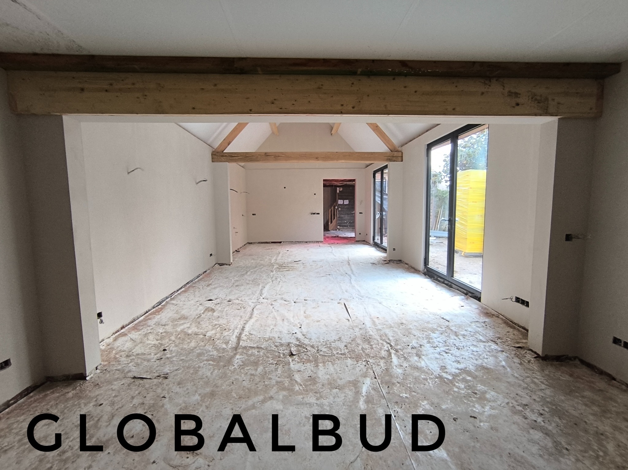 Wnętrze w stanie deweloperskim z surowymi ścianami i drewnianymi belkami stropowymi, widoczne instalacje elektryczne, duże okna tarasowe z widokiem na zewnątrz.