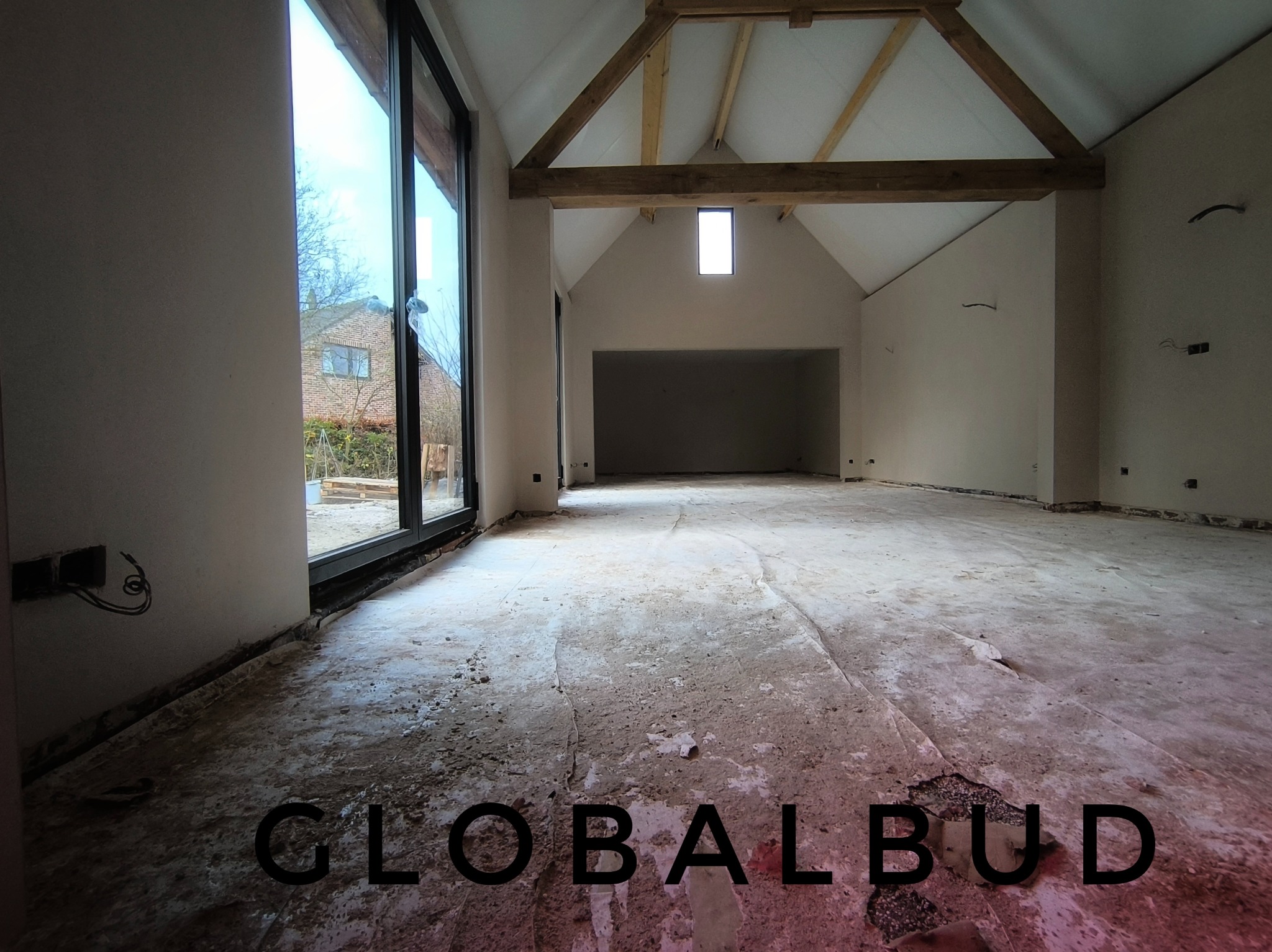 Surowe wnętrze domu w trakcie remontu: widoczne niedoskonałości podłogi, świeżo otynkowane ściany z wystającymi przewodami elektrycznymi, drewniane belki stropowe i duże okno z widokiem na budynek...