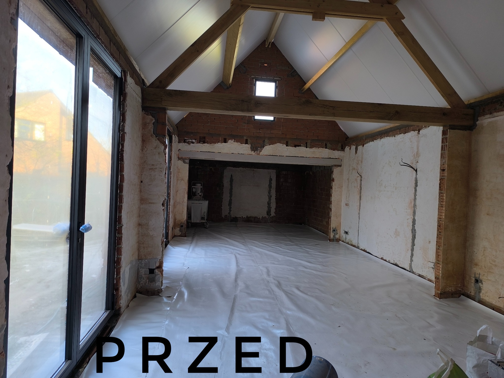 Wnętrze w stanie surowym przed tynkowaniem: widoczne cegły, drewniane belki stropowe, okno, podłoga zabezpieczona folią, napis PRZED na pierwszym planie.