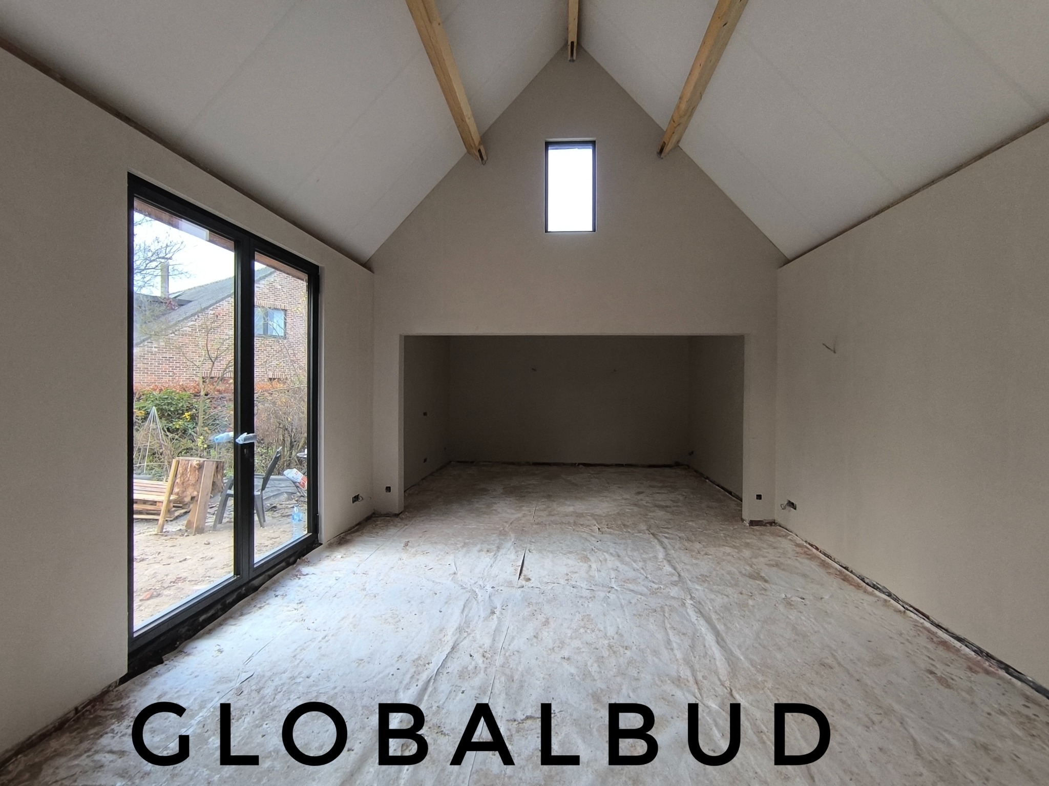 Wnętrze surowego poddasza z oknem na szczycie, po nałożeniu gładzi, z widocznymi drewnianymi belkami konstrukcyjnymi i folią ochronną na podłodze, duże okno tarasowe z widokiem na ogród.