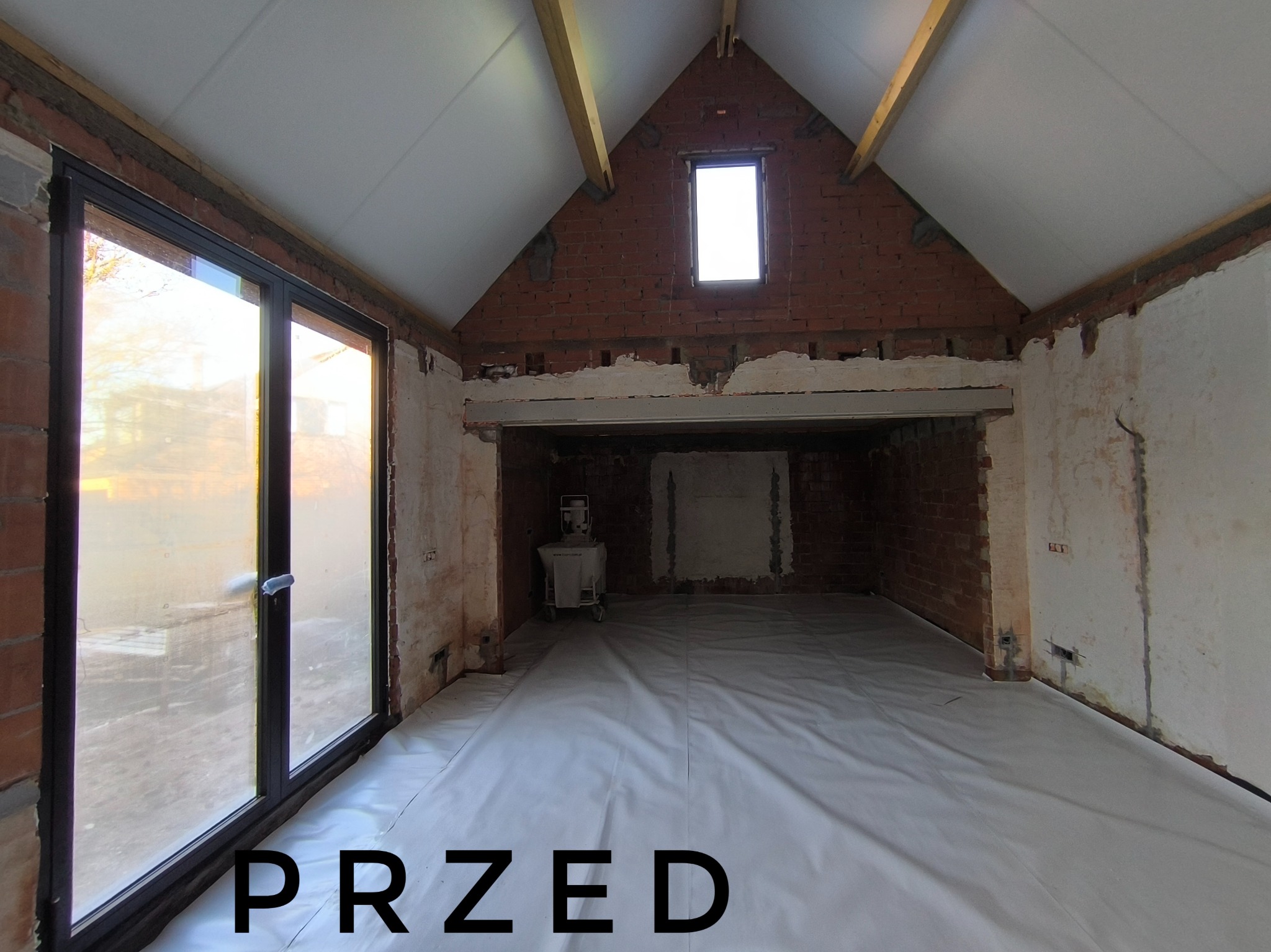 Wnętrze w trakcie remontu: surowe ściany z cegły i fragmenty tynku, okno dachowe, duże okno przesuwne, podłoga zabezpieczona folią, widoczny napis 'PRZED'.
