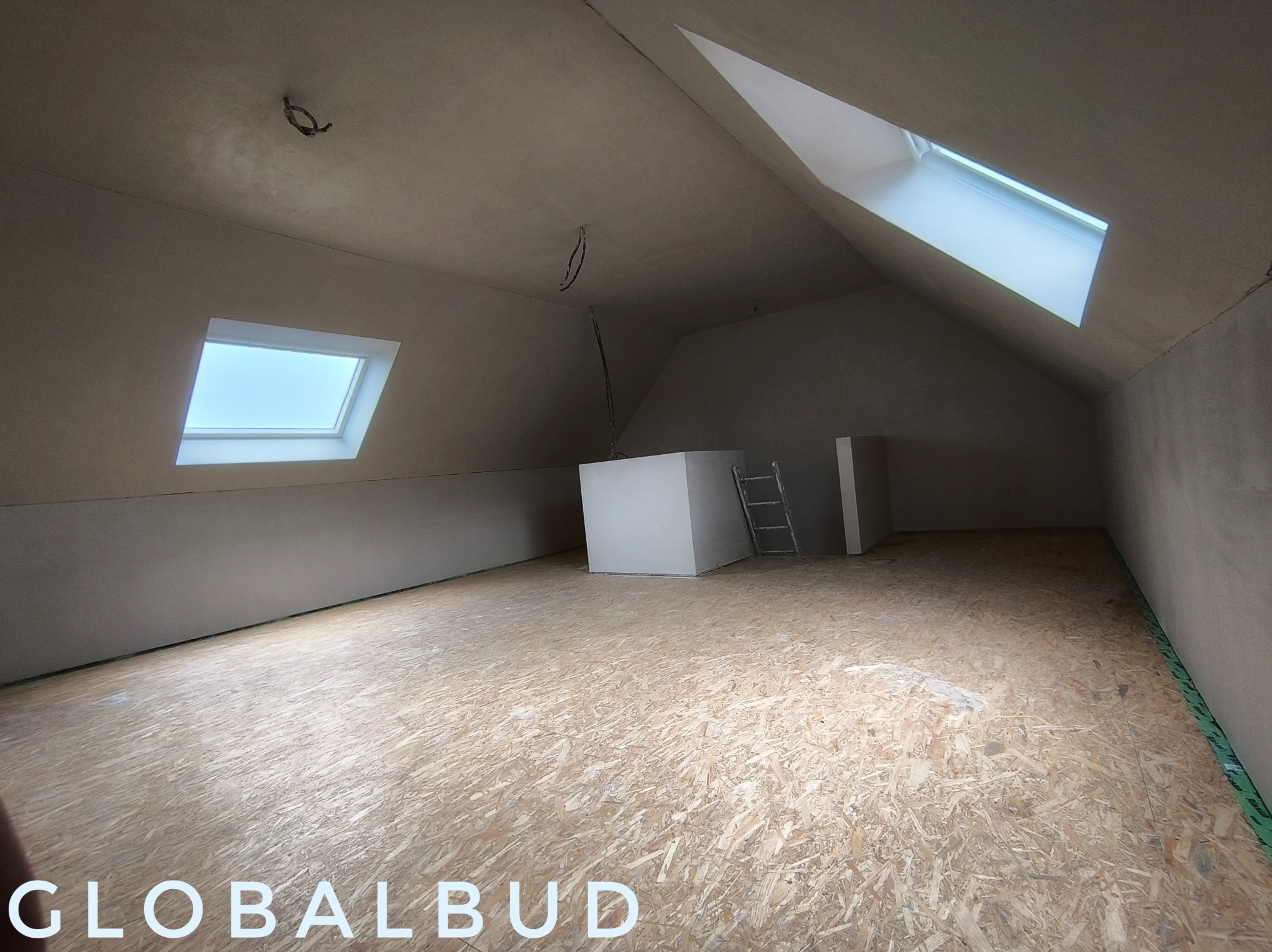 Poddasze z oknami dachowymi po pracach wykończeniowych, widoczna szara powierzchnia ścian i sufitu, drewniana podłoga OSB, drabina oparta o ścianę, widoczne kable wystające z sufitu.