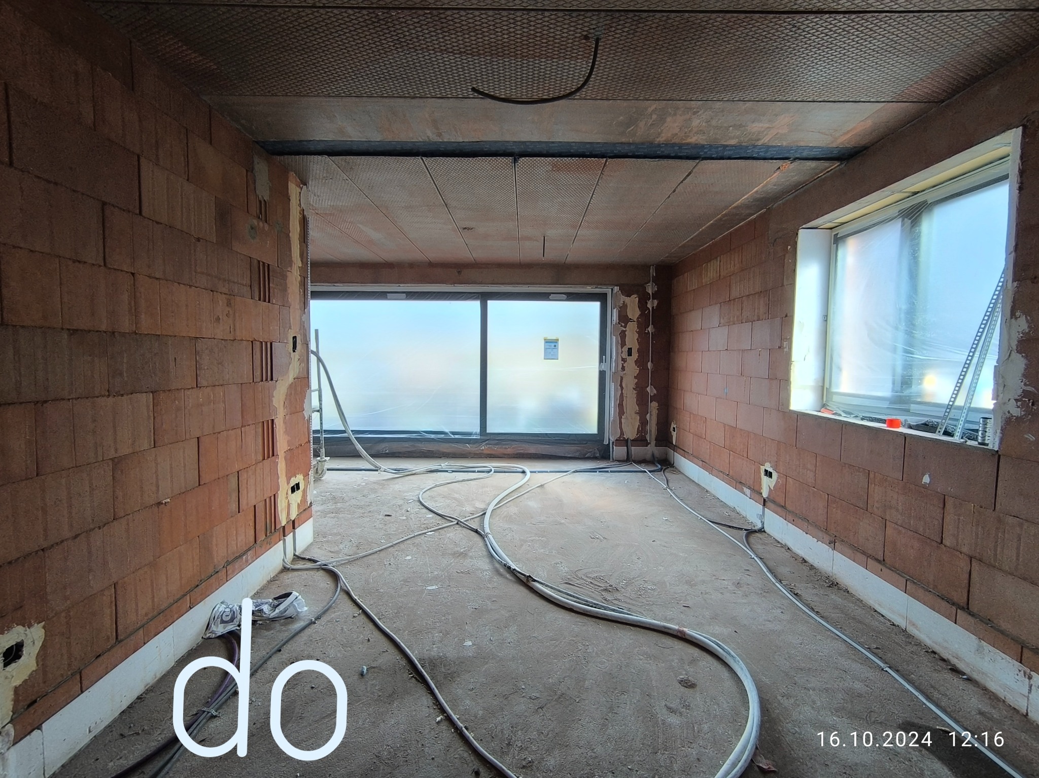 Surowy stan wnętrza z ceglanymi ścianami, przygotowane do prac wykończeniowych, widoczne przewody na podłodze i okno zabezpieczone folią.