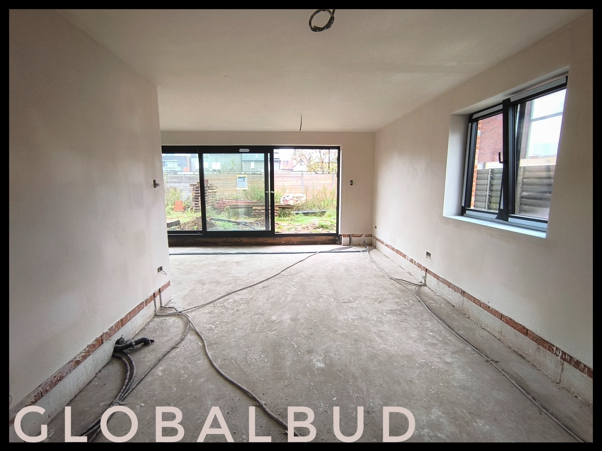 Pokój w trakcie prac wykończeniowych: świeżo nałożone tynki na ścianach, widoczne przewody na podłodze, okno i drzwi balkonowe z widokiem na ogród.