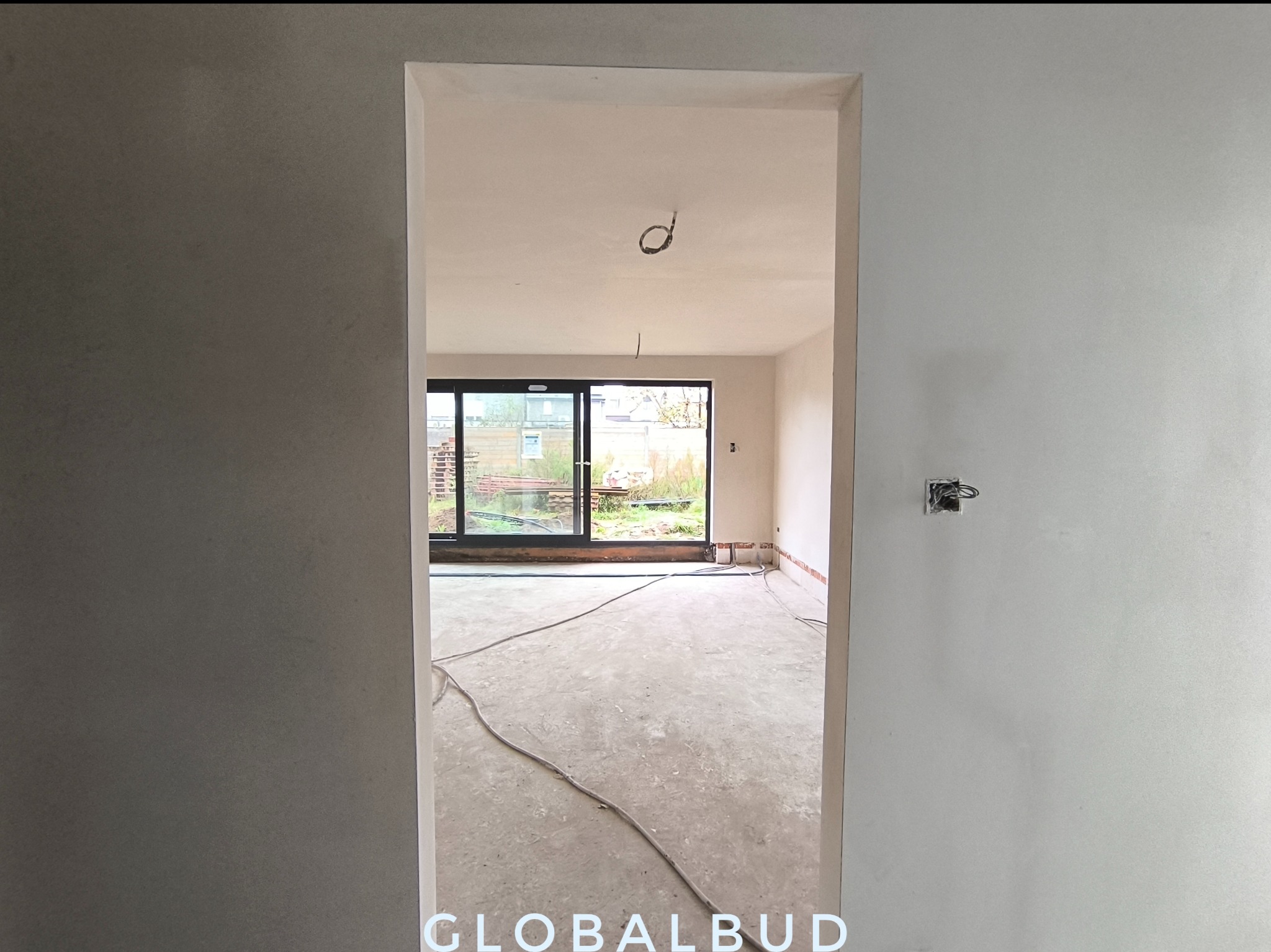 Surowe wnętrze z widoczną strukturą świeżo nałożonego tynku na ścianach, przygotowane do dalszych prac wykończeniowych, z przewodami elektrycznymi na podłodze i widokiem na ogród przez przesuwne okno.