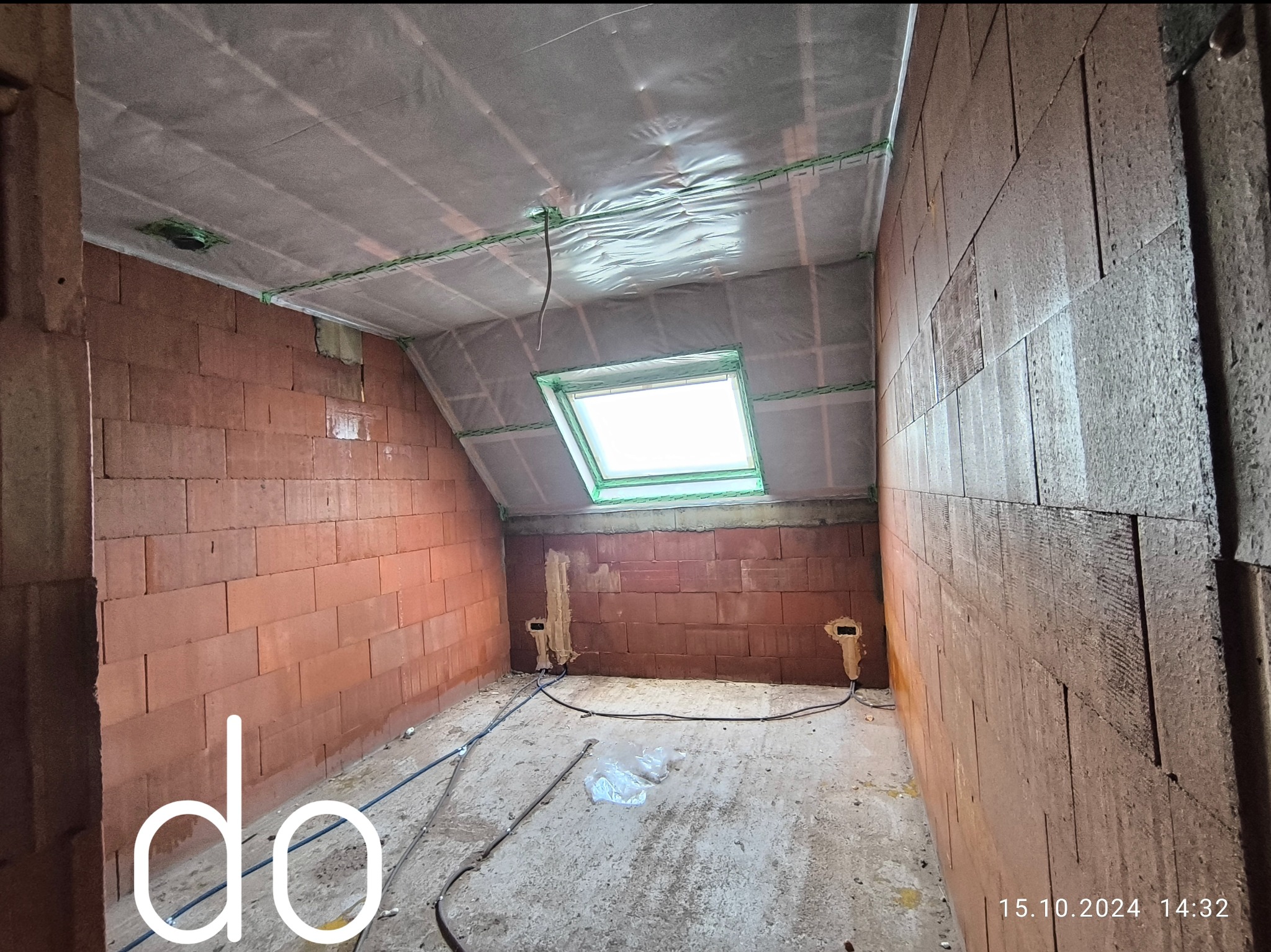 Poddasze w trakcie adaptacji: ściany z czerwonej cegły, folia paroizolacyjna na skośnym suficie, okno dachowe, widoczne przewody elektryczne i przygotowane punkty instalacyjne w ścianach.