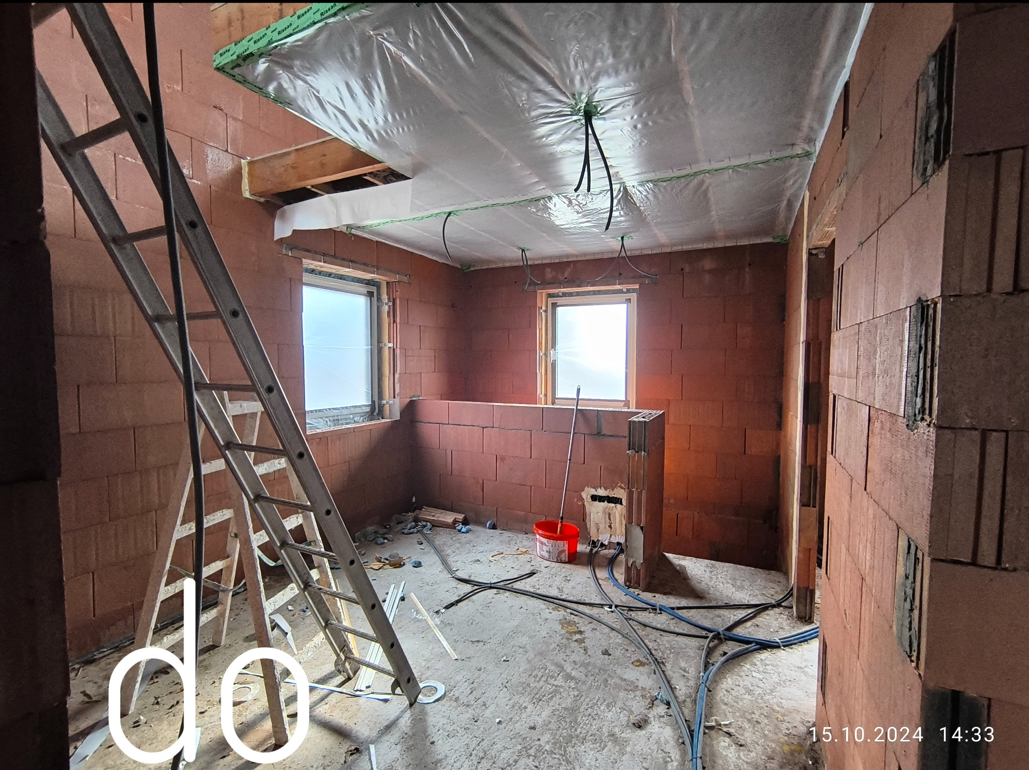 Wnętrze w trakcie budowy z czerwoną cegłą, widoczne okna, drabina oparta o ścianę, folia na suficie, kable i narzędzia na podłodze