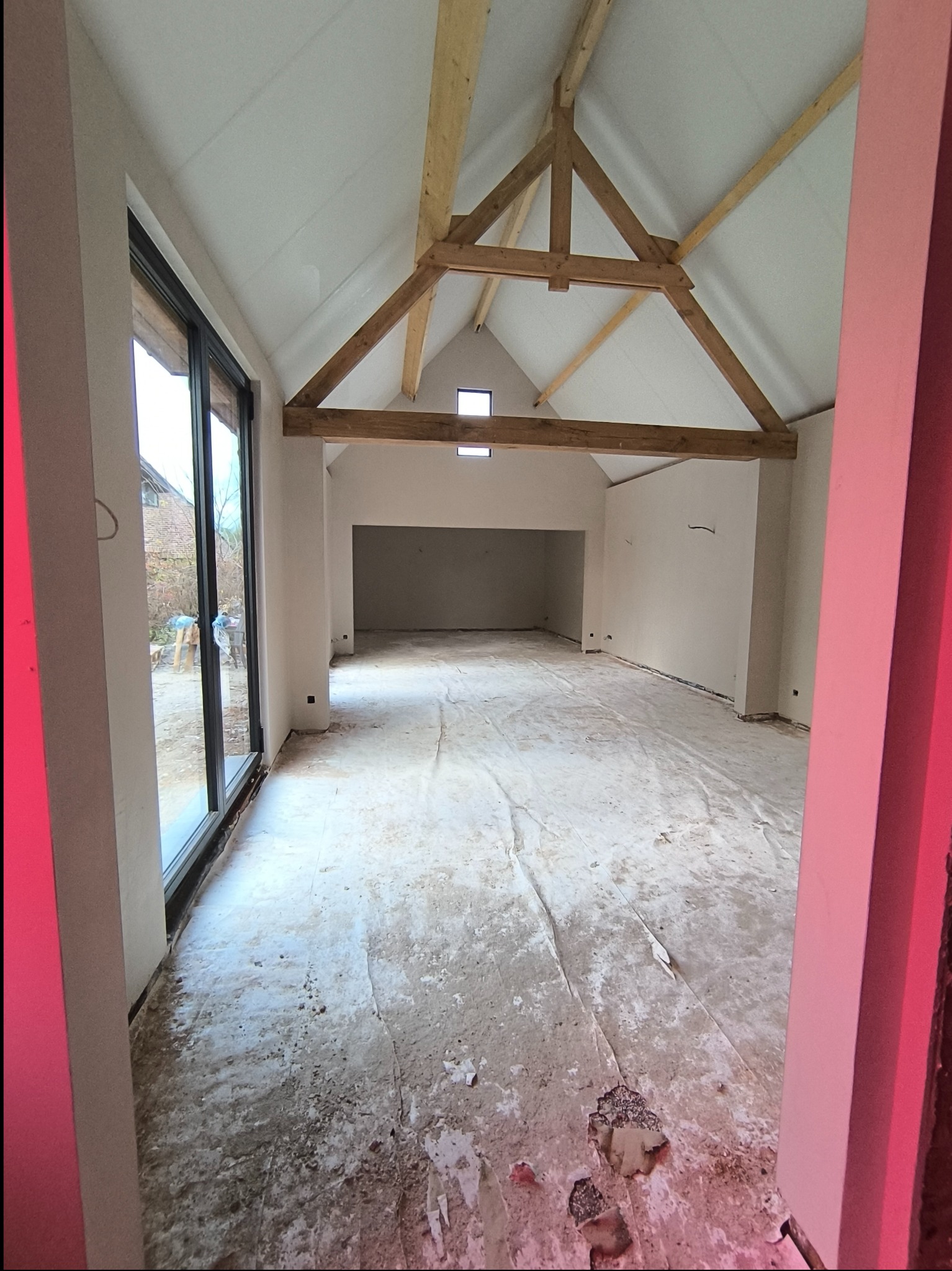 Wnętrze domu w trakcie prac wykończeniowych, widoczne drewniane elementy konstrukcji dachu, surowe ściany, duże okno i zabezpieczona folią podłoga.