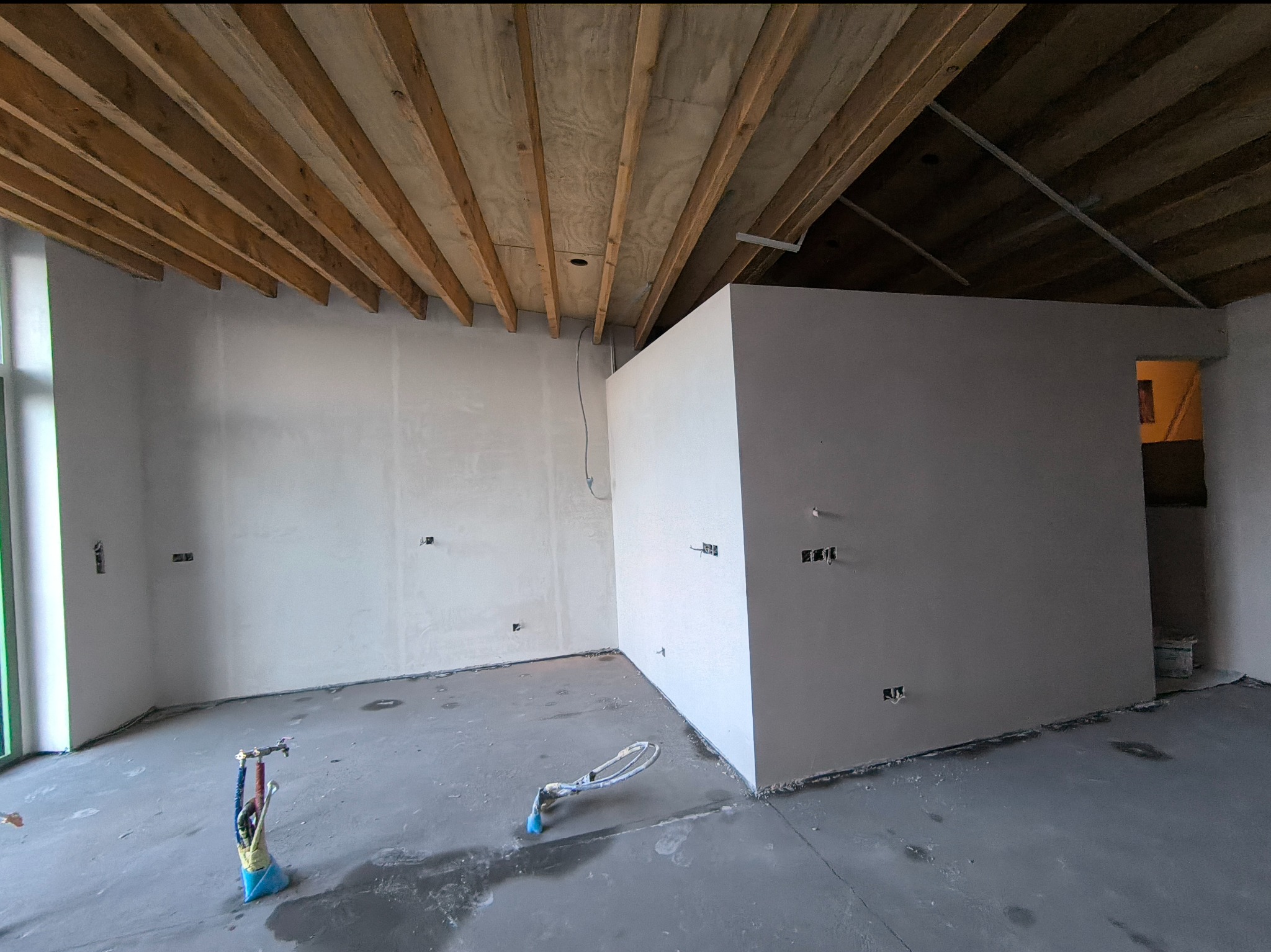 Surowe wnętrze w trakcie prac wykończeniowych: ściany pokryte świeżym tynkiem, widoczne przewody elektryczne, drewniany strop i narzędzia do tynkowania na podłodze.