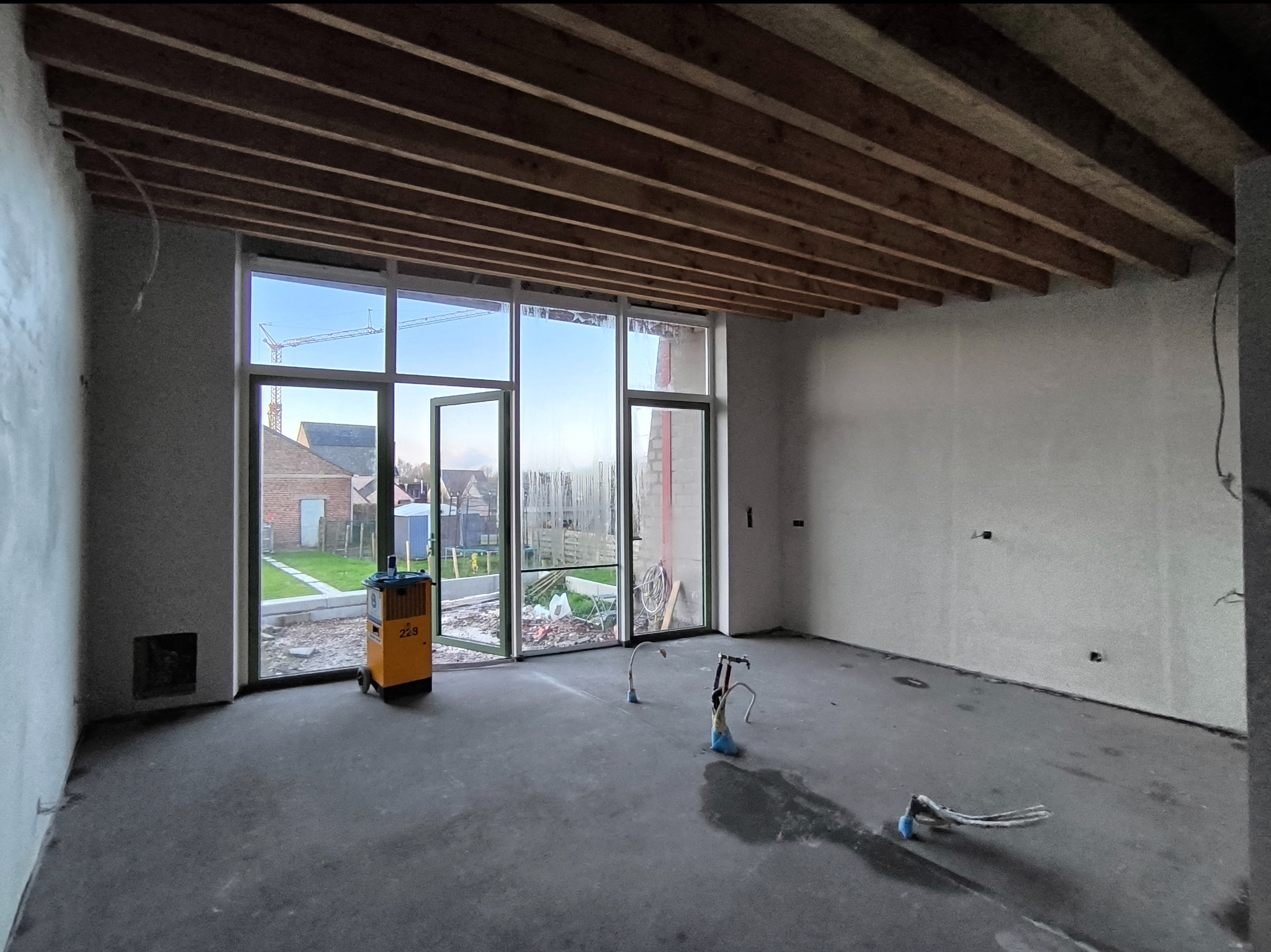 Wnętrze w stanie surowym z widocznymi śladami po instalacjach, sufit z drewnianych belek, duże okno z widokiem na ogród i dźwig budowlany w oddali, żółty osuszacz powietrza przy oknie.