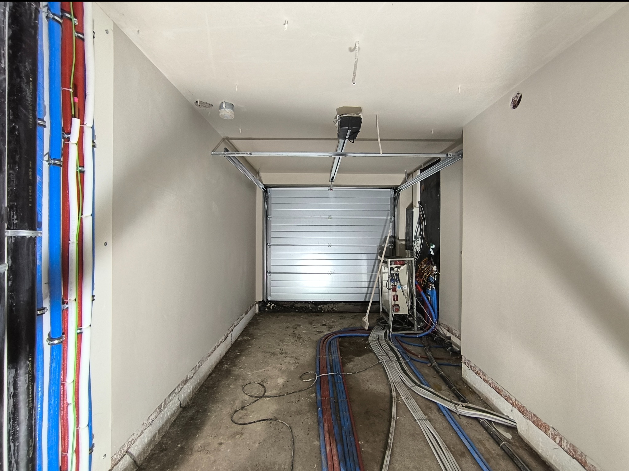 Wnętrze garażu w trakcie prac wykończeniowych: świeżo otynkowane ściany, instalacje elektryczne i hydrauliczne prowadzone wzdłuż ścian, widoczny mechanizm bramy garażowej.