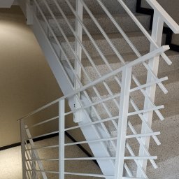 DAW-TECH - Białe, metalowe schody z minimalistyczną balustradą wewnątrz budynku, widok z góry na dół, schody wykończone lastryko.