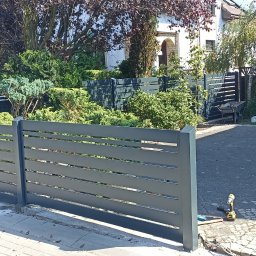 DAW-TECH - Nowoczesne, poziome ogrodzenie panelowe w kolorze szarym, z widocznymi narzędziami montażowymi na kostce brukowej, otoczone zielenią i zabudową jednorodzinną.