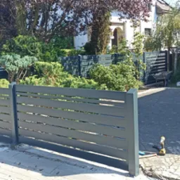 Nowoczesne, poziome ogrodzenie panelowe w kolorze szarym, z widocznymi narzędziami montażowymi na kostce brukowej, otoczone zielenią i zabudową jednorodzinną.