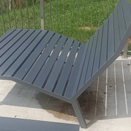 DAW-TECH - Nowoczesny, szary leżak ogrodowy z metalową ramą i drewnianymi listwami, rzucający cienie na betonowe podłoże, z widocznym fragmentem metalowej balustrady w tle i fragmentem osoby w kolorowych...