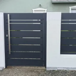 Nowoczesna, antracytowa brama wjazdowa z poziomymi panelami, osadzona między białymi, murowanymi słupkami z domofonem, na tle zielonego domu.