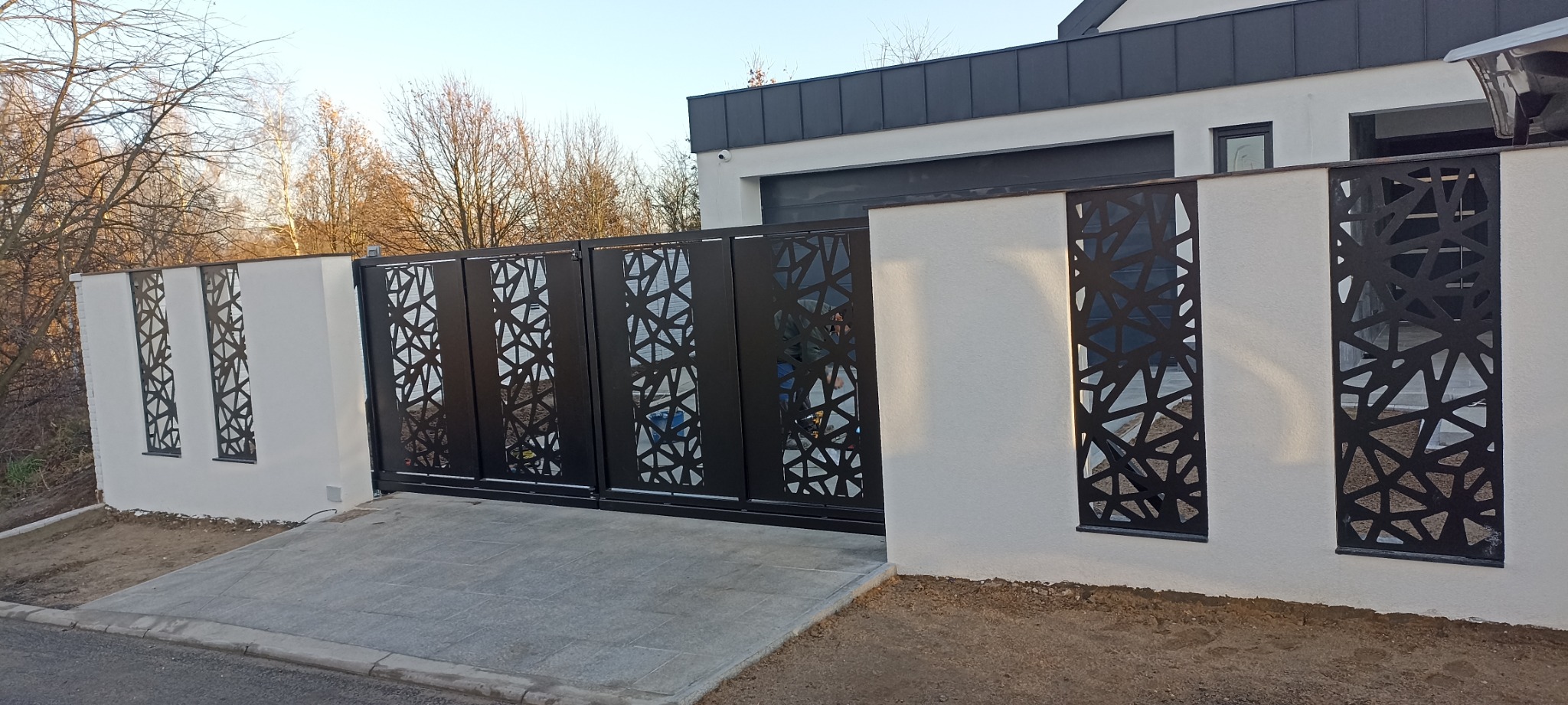 Nowoczesna brama wjazdowa z metalowymi panelami o ażurowym wzorze, wkomponowana w biały mur z dekoracyjnymi wstawkami o identycznym motywie, na tle domu z garażem.