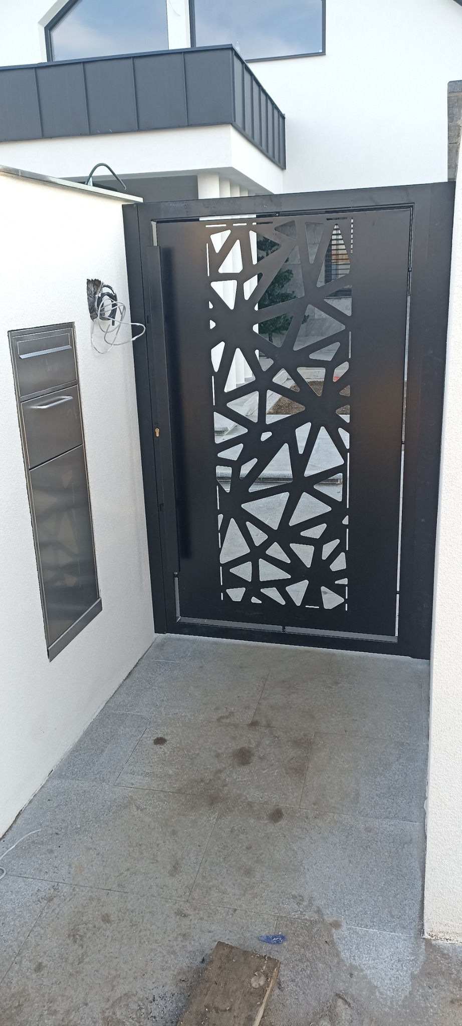 Nowoczesna, czarna brama wjazdowa z geometrycznym wzorem wyciętym laserowo, zamontowana przy minimalistycznym budynku z szarą skrzynką na listy.